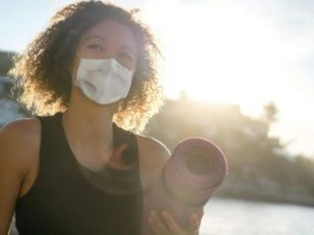 Beautiful young woman holding yoga mat and wearing mask in the pandemic time